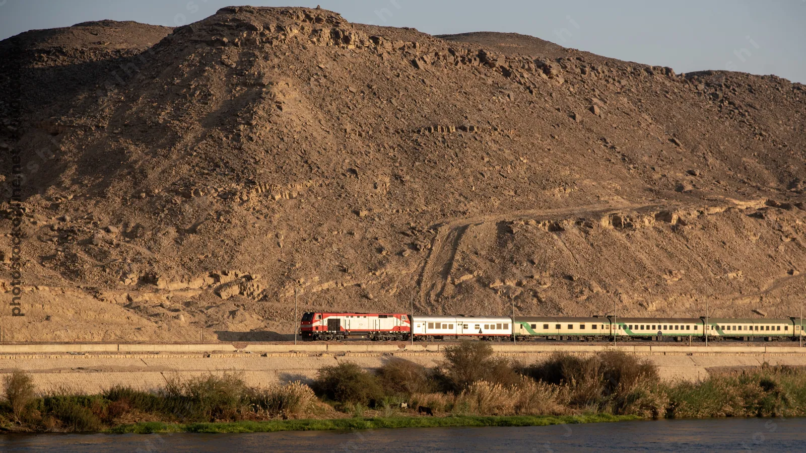 Train traveling between the Nile River and a desert mountain near Aswan, Egypt - a stunning landscape of water and rugged terrain.