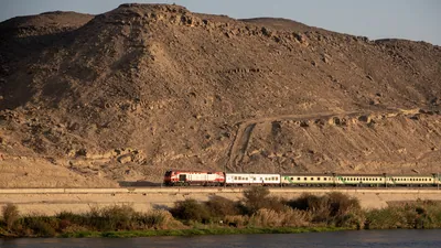 Train traveling between the Nile River and a desert mountain near Aswan, Egypt - a stunning landscape of water and rugged terrain.