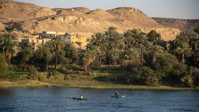 Fishing boats on the Nile River near Aswan, Egypt, with palm-lined banks and brick houses nestled on the mountain slope in the background.