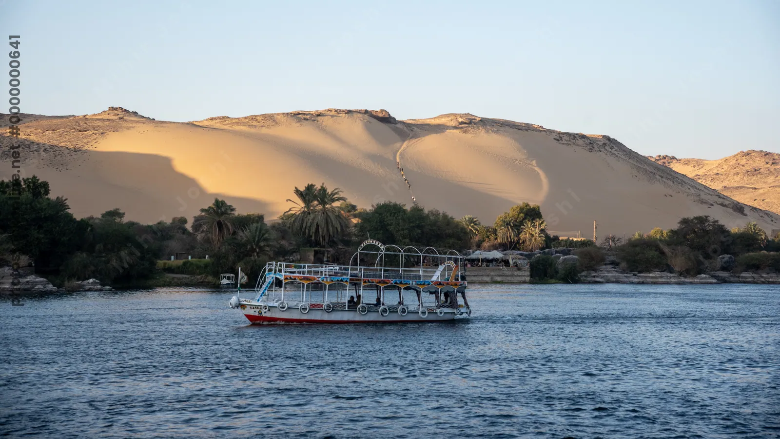 Aswan, Egypt - Feb 7, 2025: Tour boats cruising the Nile River at Aswan, Egypt, with a huge sand dune forming a dramatic desert backdrop.