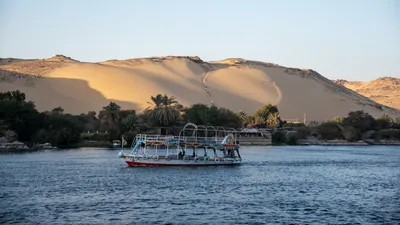 Aswan, Egypt - Feb 7, 2025: Tour boats cruising the Nile River at Aswan, Egypt, with a huge sand dune forming a dramatic desert backdrop.