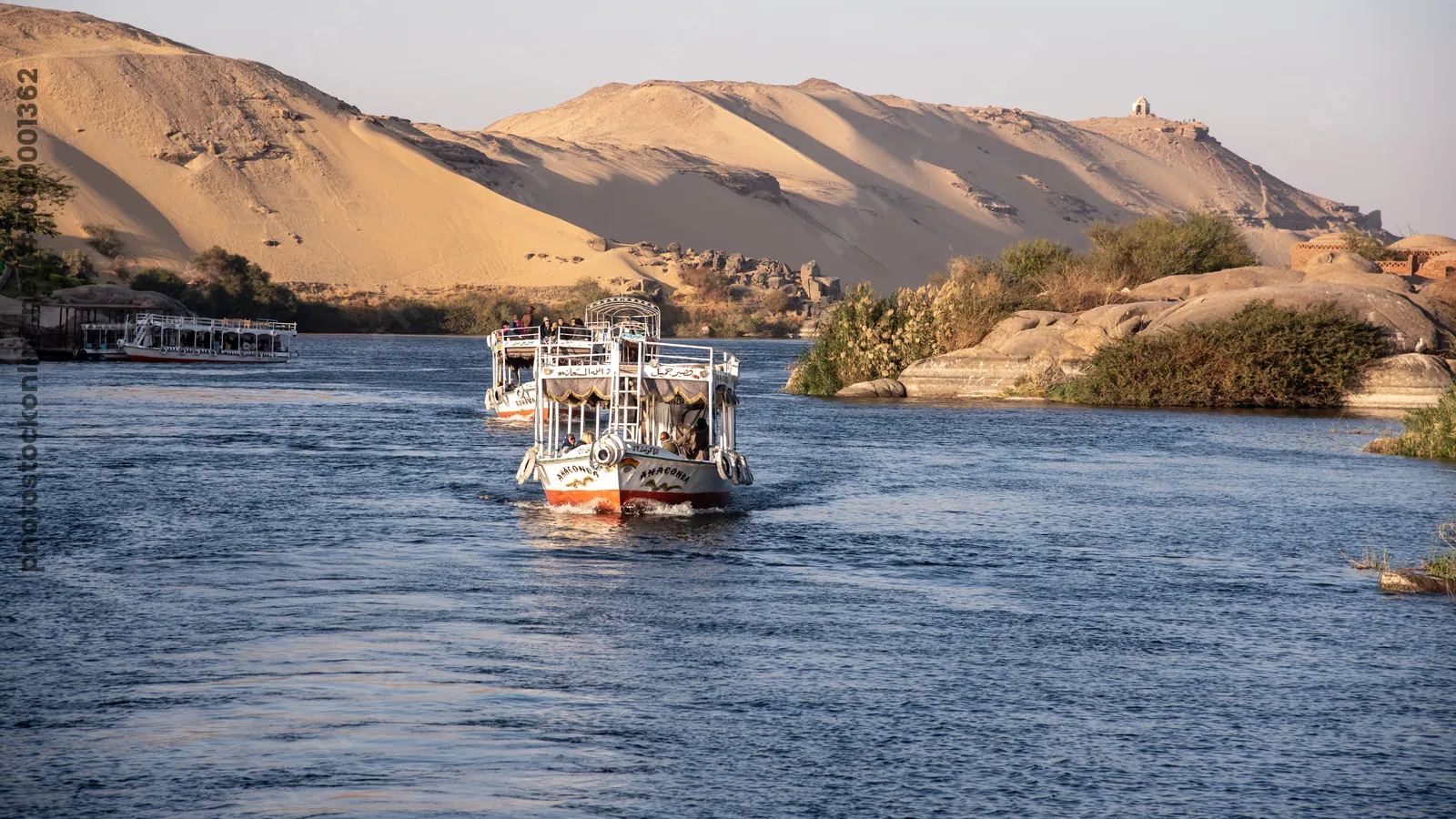 Aswan, Egypt - Feb 7, 2025: Tour boats cruising the Nile River at Aswan, Egypt, with a huge sand dune forming a dramatic desert backdrop.