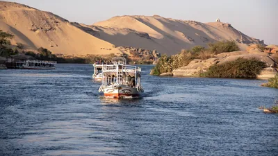 Aswan, Egypt - Feb 7, 2025: Tour boats cruising the Nile River at Aswan, Egypt, with a huge sand dune forming a dramatic desert backdrop.