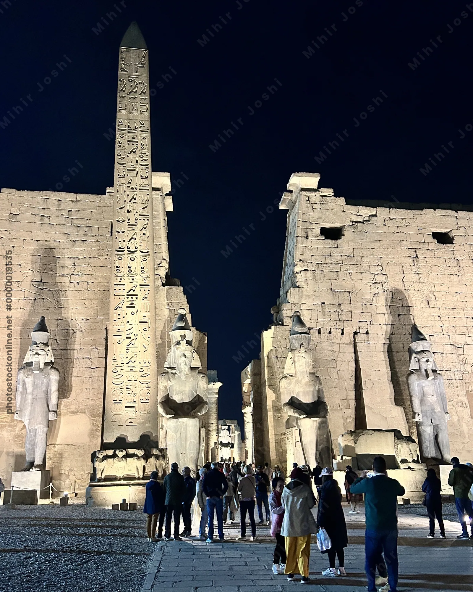 Luxor, Egypt - Feb 9, 2025: Illuminated entrance of Luxor Temple at night with colossal statues and a standing obelisk, with visitors in the foreground.