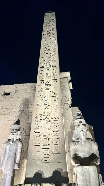 Luxor, Egypt - Feb 9, 2025: Night view of the illuminated obelisk and colossal statues at the entrance pylon of Luxor Temple in Egypt, with hieroglyphs carved in stone.