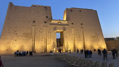 Edfu, Egypt - Feb 9, 2025: Dawn view of the illuminated entrance pylons of the Temple of Edfu in Egypt, with monumental relief carvings and visitors in the foreground.