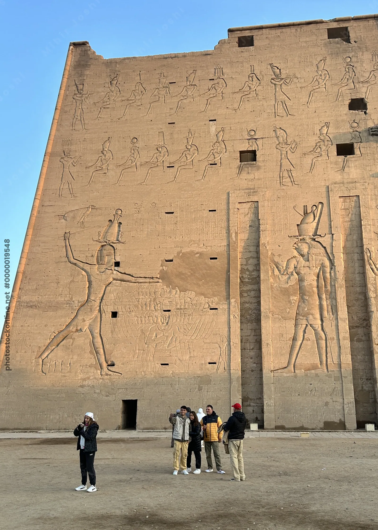 Edfu, Egypt - Feb 9, 2025: Visitors stand before the monumental pylon wall of the Temple of Edfu in Egypt, decorated with ancient relief carvings of pharaohs and deities.