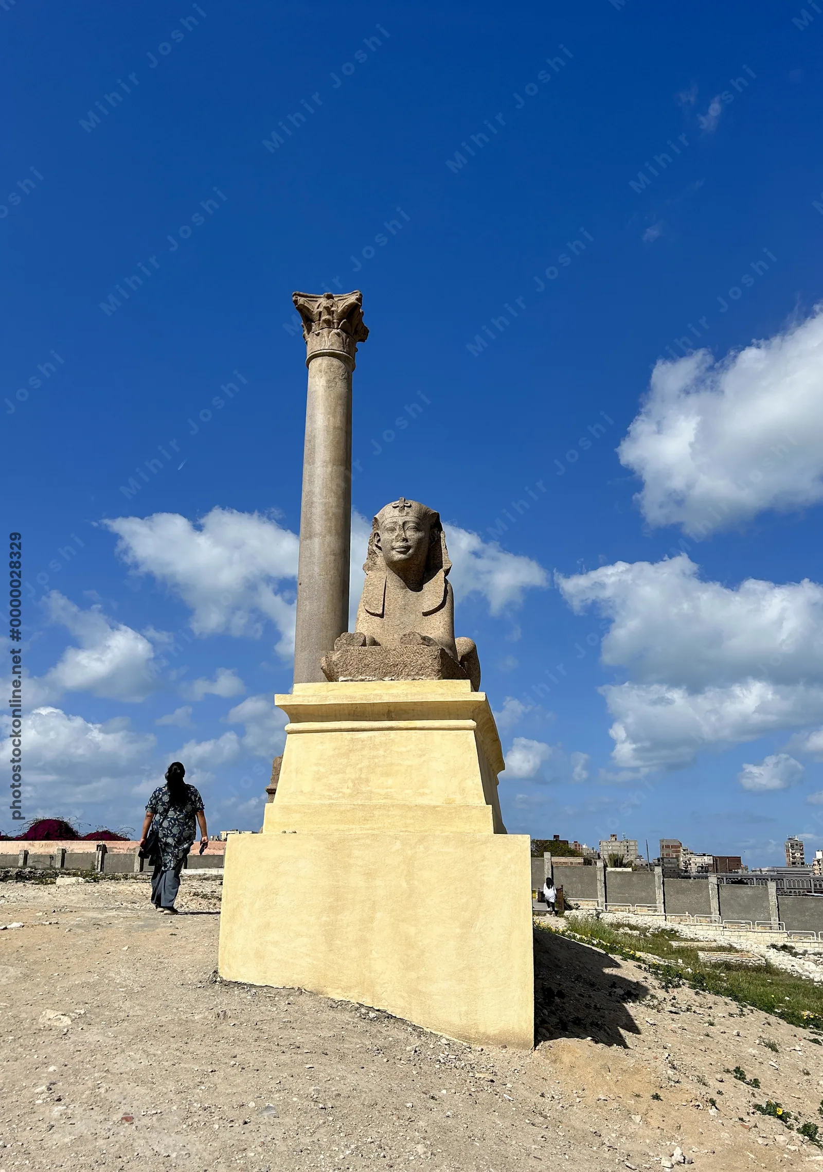 Alexandria, Egypt - Feb 13, 2025: Granite sphinx statue with Pompey's Pillar at the ancient Serapeum of Alexandria in Egypt under a blue sky.