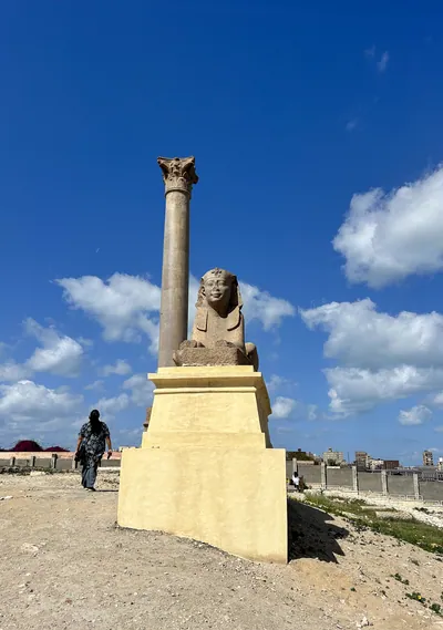 Alexandria, Egypt - Feb 13, 2025: Granite sphinx statue with Pompey's Pillar at the ancient Serapeum of Alexandria in Egypt under a blue sky.