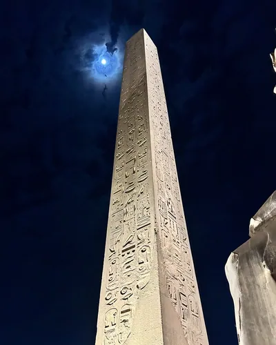 Luxor, Egypt - Feb 9, 2025: Night view of the illuminated obelisk of Pharaoh Ramses II at Luxor Temple in Egypt, with the moon glowing through clouds