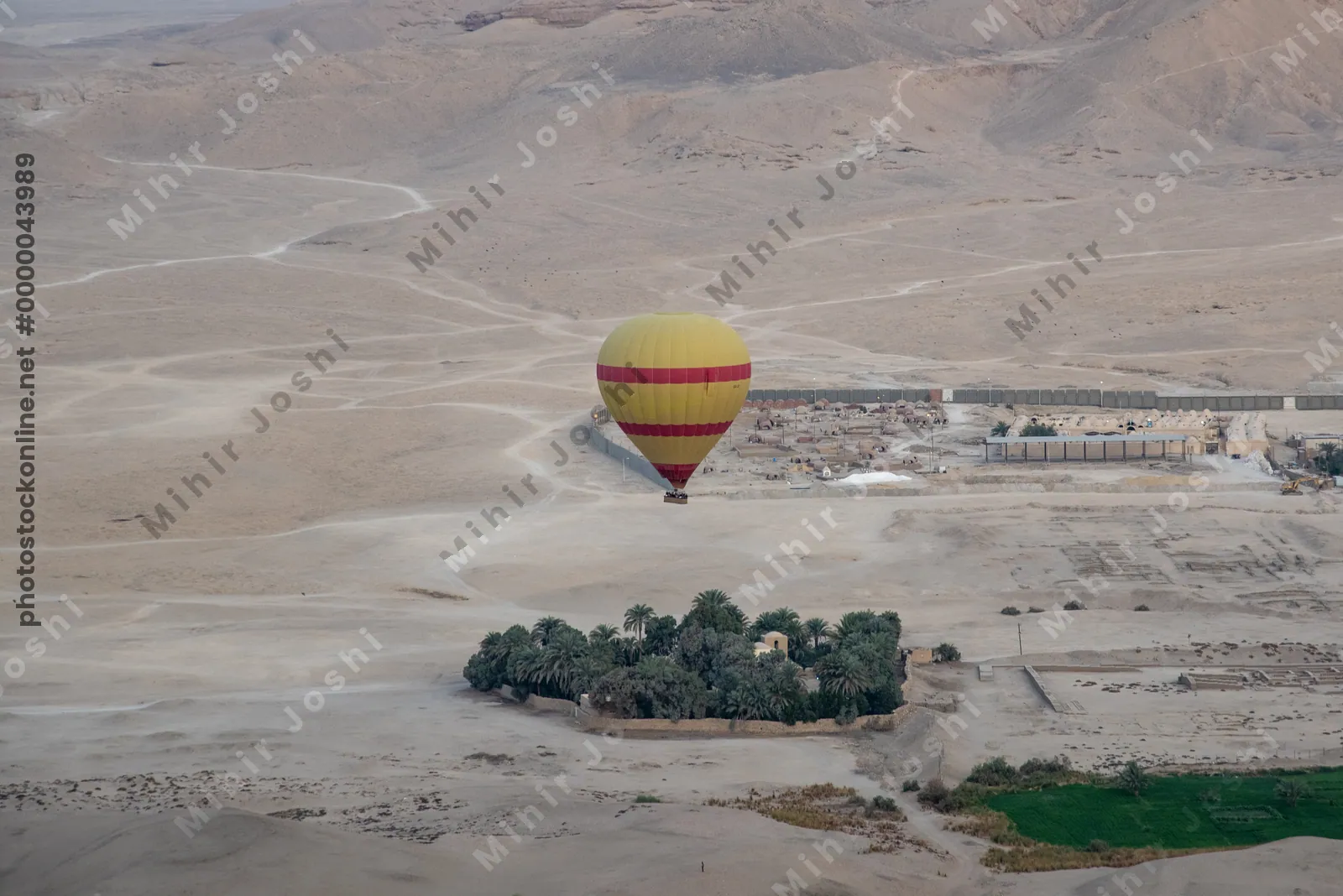 A colorful hot air balloon floats above a green oasis surrounded by desert near Luxor, Egypt, during sunrise