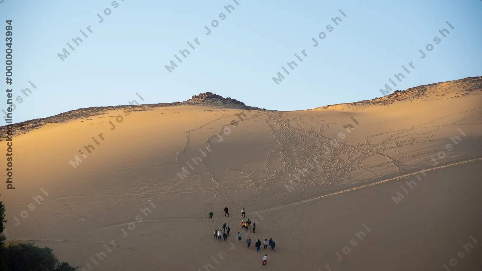 Tourists climbing a massive sand dune near the Nile River at Aswan, Egypt, during sunset with dramatic shadows on the dunes.