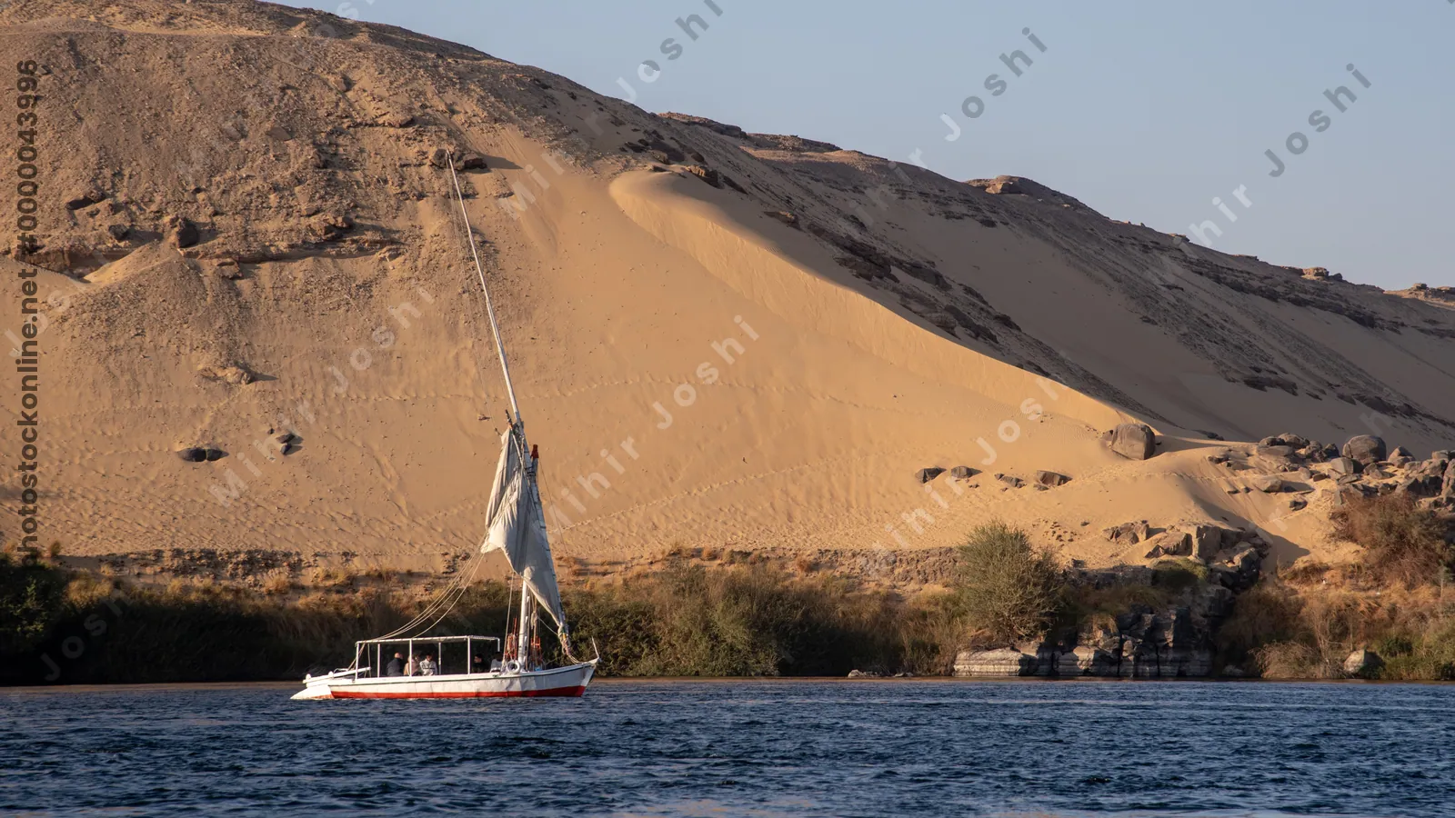 Local Egyptians sailing a felucca on the Nile River at Aswan. First Cataract with large sand dunes in the background. Authentic cultural travel scene.