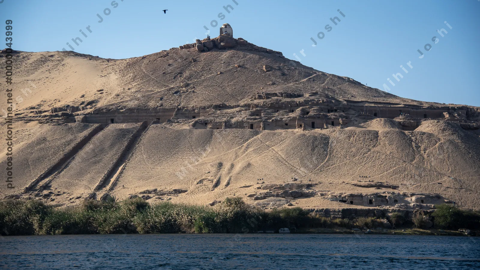 Ancient rock-cut Tombs of the Nobles on the hillside across the Nile River from Aswan, Egypt, seen from the riverbank.