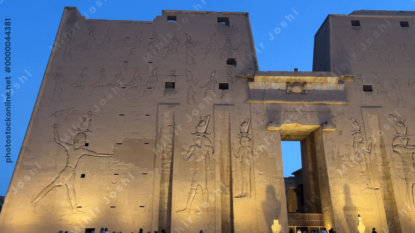 Edfu, Egypt - Feb 9, 2025: Dawn view of the illuminated entrance pylons of the Temple of Edfu in Egypt, with monumental relief carvings and visitors in the foreground.