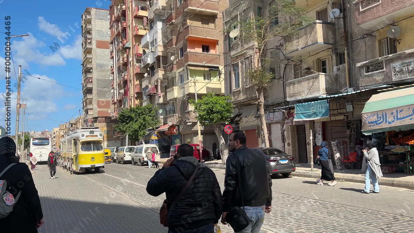 Alexandria, Egypt - Feb 13, 2025: Yellow tram running through a busy street with pedestrians, cars, and market stalls in the city center.