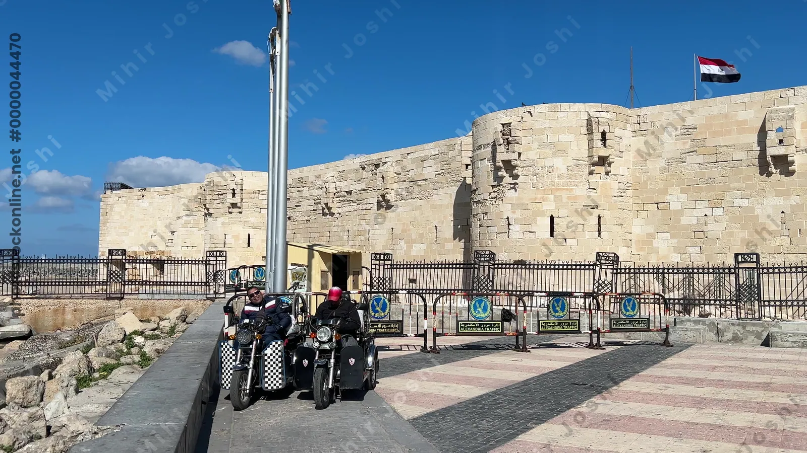 Alexandria, Egypt - Feb 13, 2025: Side view of the Citadel of Qaitbay fortress with motorcycles parked nearby and fishing boats along the Mediterranean coast.