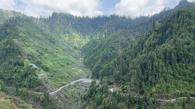 View of terraced hills, forested slopes and winding mountain road with scattered houses in Janjehli valley, Mandi district, Himachal Pradesh, India.